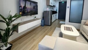 Una sala de estar moderna con un televisor de pantalla plana, un sofá, una mesa de centro y una zona de cocina con nevera y armarios.
