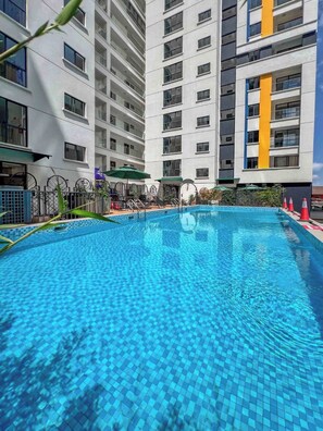 Uma piscina com piso em azulejo, cercada por uma grade, e adjacente a um edifício com várias andares e varandas.