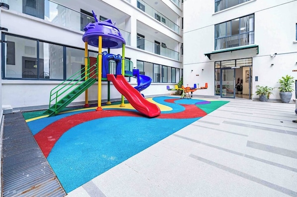 Um parquinho com escorregadores e estruturas de escalada, cercado por um edifício com grandes janelas e telhado plano.