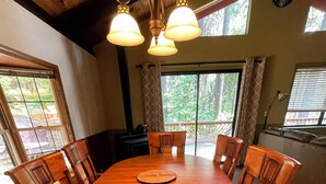 A wooden dining table with six chairs, a clock on the wall, and large windows with curtains. - Cozy 2-bedroom cabin in Pollock Pines, Apple Hill/Gold Country (Pollock Pines)