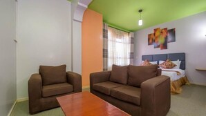 A room with two brown sofas, a wooden coffee table, a bed with a blue headboard, and a window with curtains. - maple Inn Hotel (Ruiru)