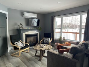 A living room with a fireplace, a television mounted above it, and a large window with a view of a snowy deck.