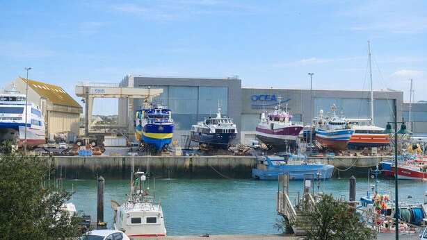 Un port de plaisance où sont amarrés divers bateaux, un grand bâtiment industriel portant l’inscription « OCEA », et un ciel dégagé.