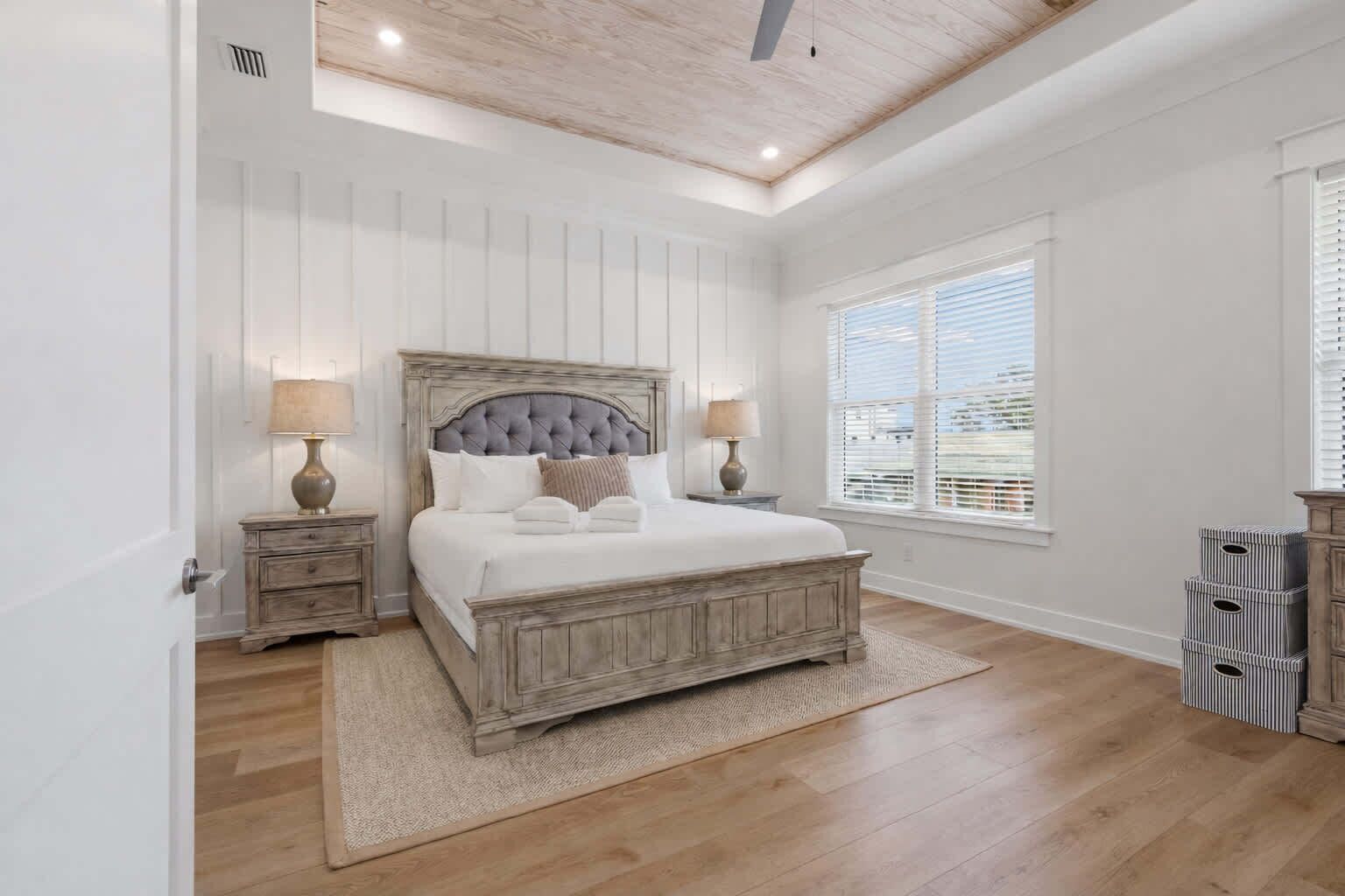 A bedroom with a large bed, two bedside tables, a ceiling fan, a window with blinds, and a wooden floor.