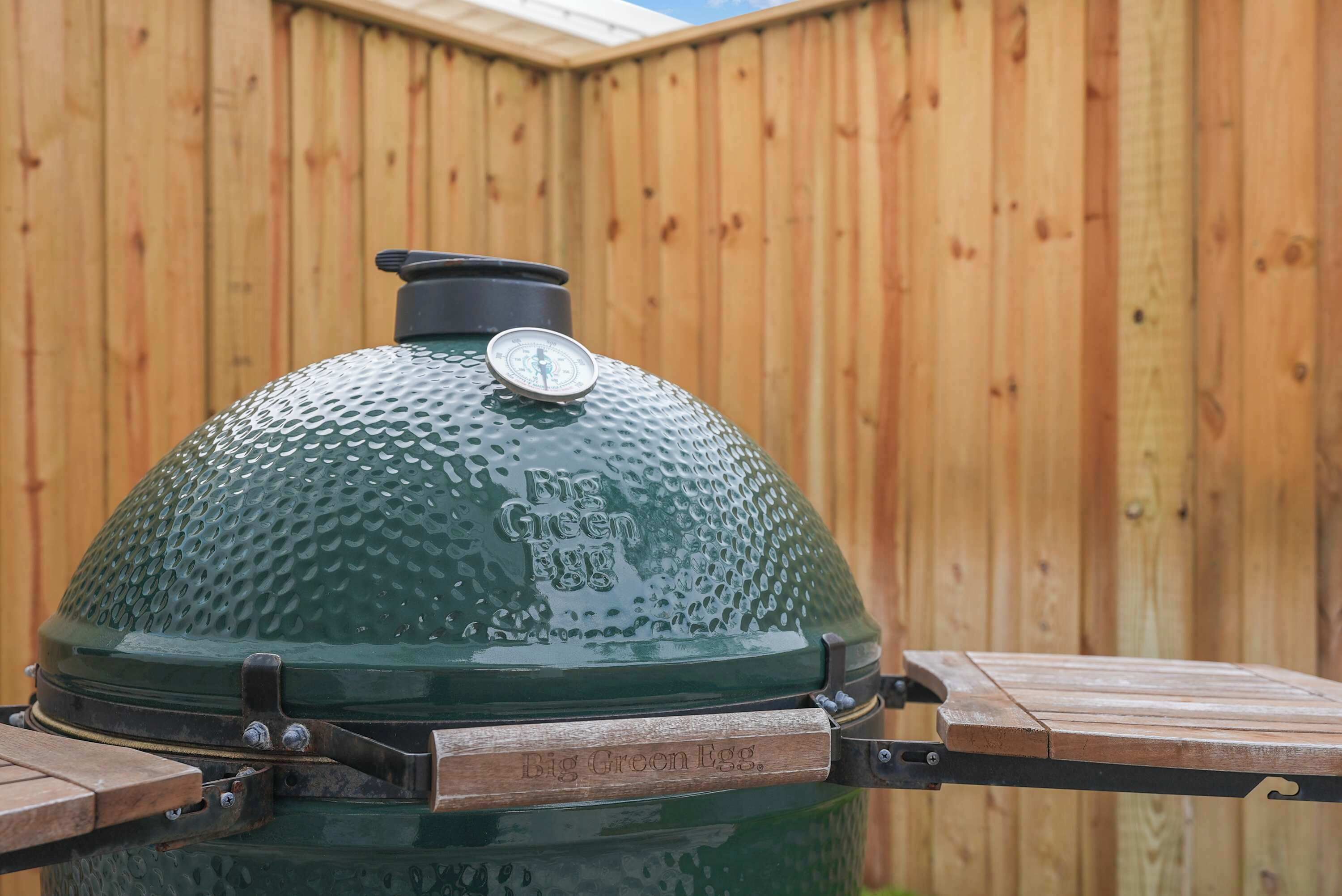 A green barbecue grill with a thermometer and a metal handle, set against a wooden fence.