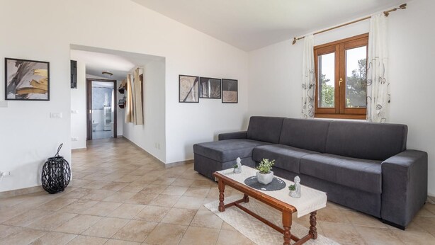 Uma sala de estar com um sofá cinzento, uma mesa de centro de madeira e uma janela com cortinas.