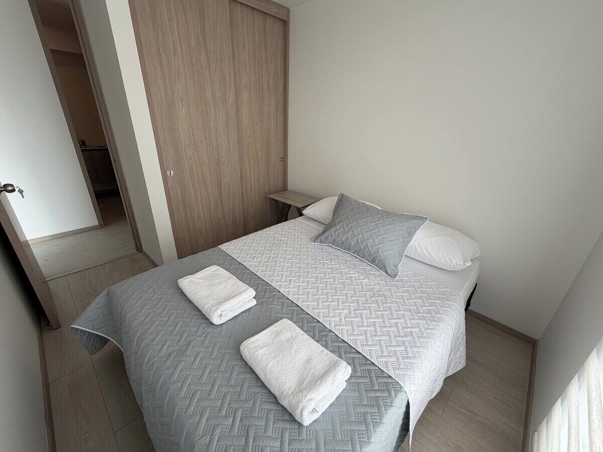 A neatly made bed with a grey and white patterned bedspread, two pillows, and two folded towels.