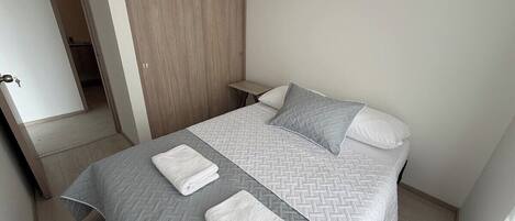 A neatly made bed with a grey and white patterned bedspread, two pillows, and two folded towels.