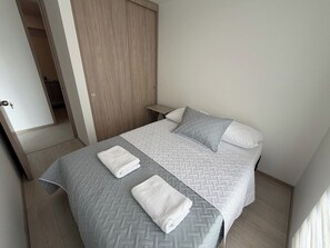 Cama bem arrumada com colcha cinza e branca estampada, dois travesseiros e duas toalhas dobradas.