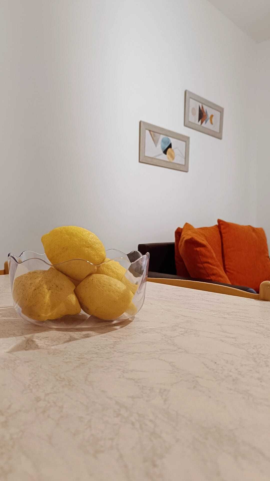 Una ciotola di vetro trasparente con limoni su una superficie in marmo, una sedia nera e un cuscino arancione in una stanza con pareti bianche e quadri appesi.