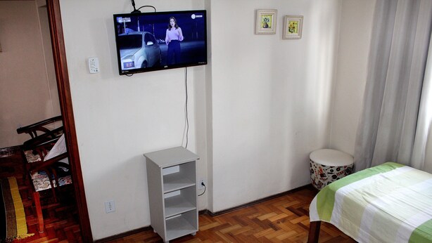 Quarto com cama, televisão fixada na parede, prateleira pequena branca e um banquinho redondo.