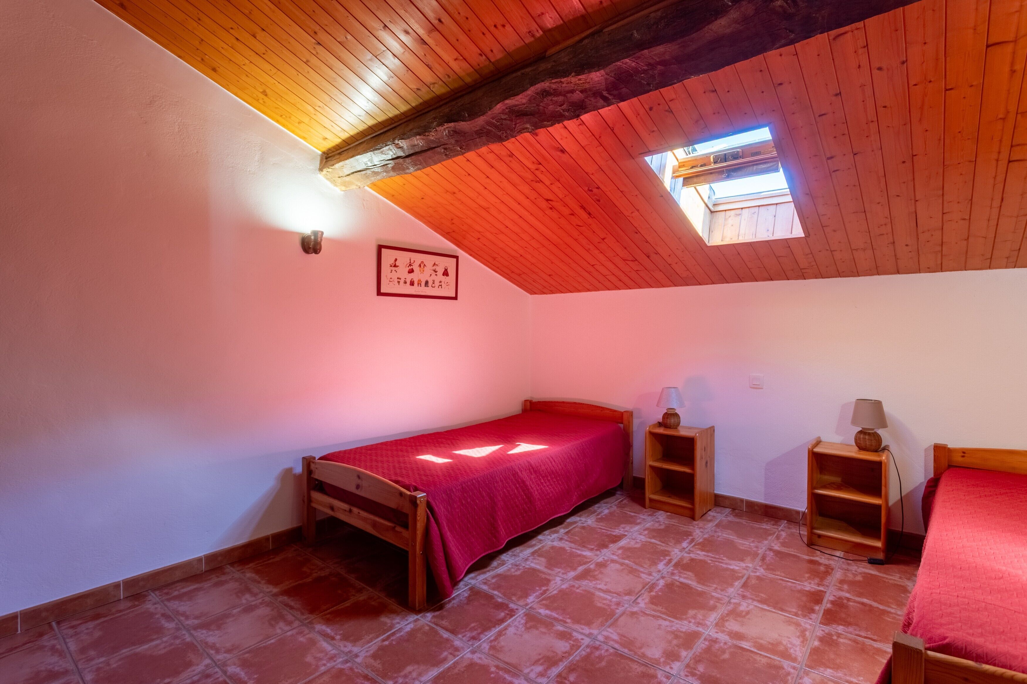 Une chambre avec un plafond en pente, un puits de lumière, un lit recouvert d’une couverture rouge, une table de chevet avec une lampe et une petite étagère en bois.