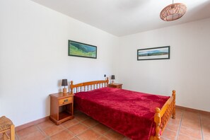 Quarto com cama, mesas de cabeceira de madeira, colcha vermelha, duas imagens penduradas na parede e luminária de teto trançada.