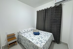 A small, neatly arranged bedroom with a bed, a small wooden shelf, and a curtain.