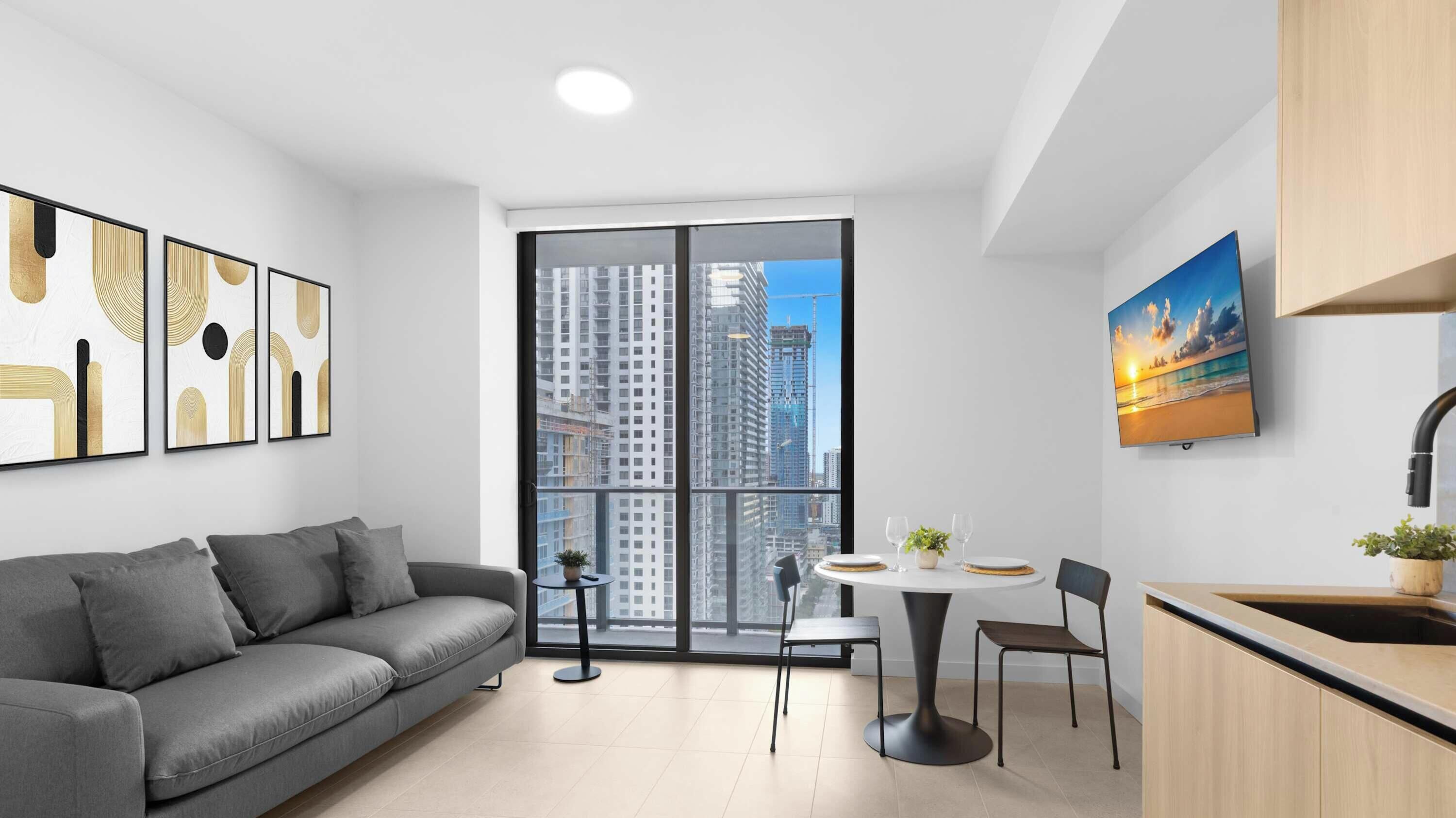 A modern apartment with a grey sofa, a dining table with chairs, a kitchen area, and large windows offering a city view.