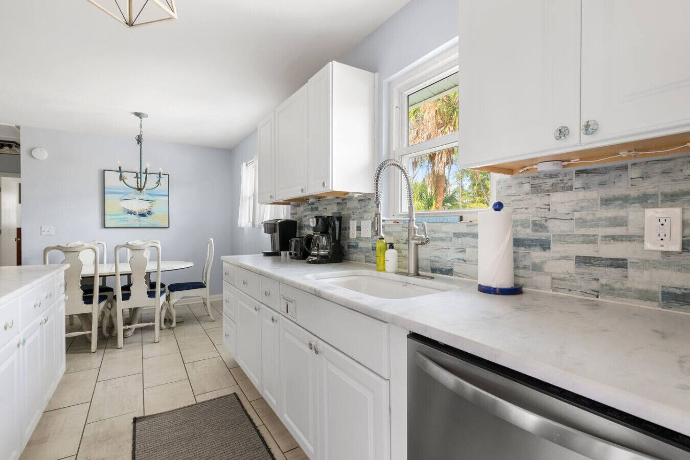 A modern kitchen with white cabinets, a built-in dishwasher, and a window with a view of palm trees.