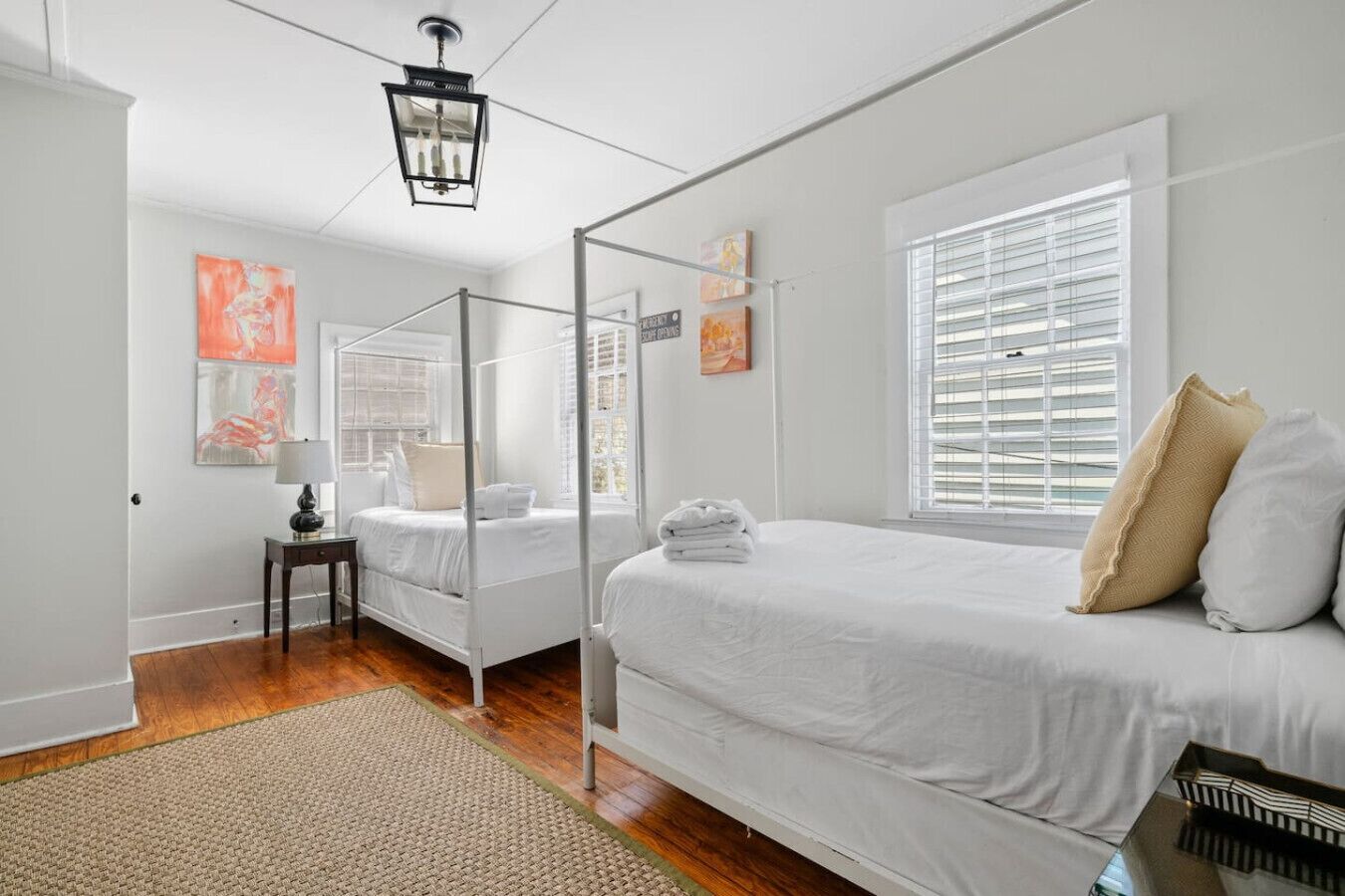 Une chambre à coucher avec un lit à baldaquin, une table de chevet, une lampe, une fenêtre avec des stores et des œuvres d’art accrochées au mur.