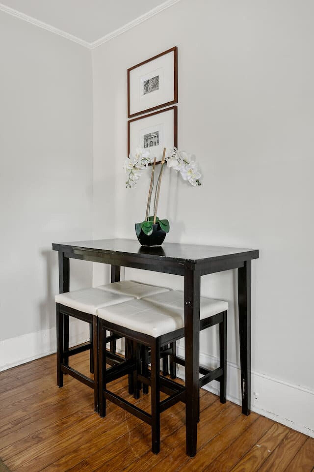 A dark wooden dining table with white cushioned chairs, a vase with flowers, and framed pictures on the wall.