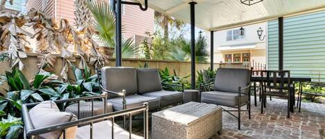 An outdoor patio with a sofa, chairs, and a table, surrounded by plants and a brick floor.