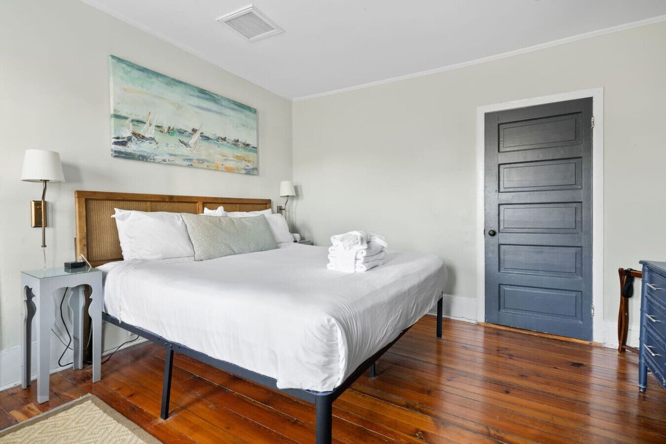 A neatly made bed with white linens, a wooden headboard, and a painting of a seascape on the wall.