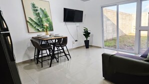 Un salon comprenant un canapé, une table à manger avec des chaises, un téléviseur fixé au mur et une porte-fenêtre coulissante donnant sur un espace extérieur.