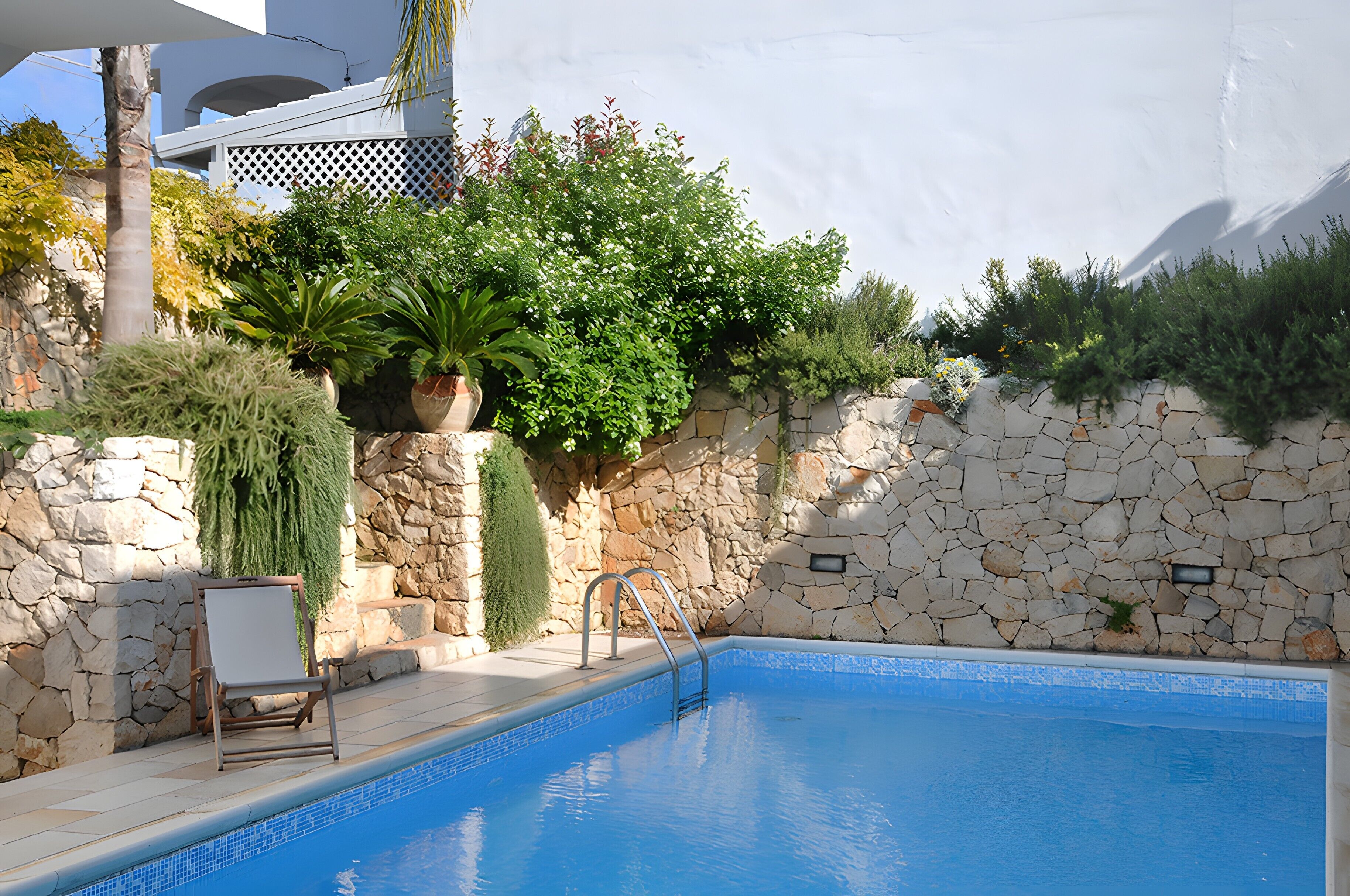 A swimming pool with a stone wall, a chair, and potted plants.