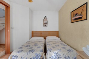 Une chambre avec deux lits, une tête de lit en bois, une étagère fixée au mur avec une maquette de bateau, et un escalier sur le côté gauche.