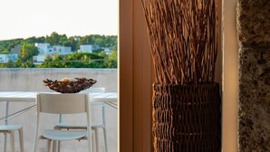 Ein Balkon mit Blick auf ein Gewässer und grüne Bepflanzung, eine Korbbrosche mit Zweigen und ein Tisch mit Stühlen.