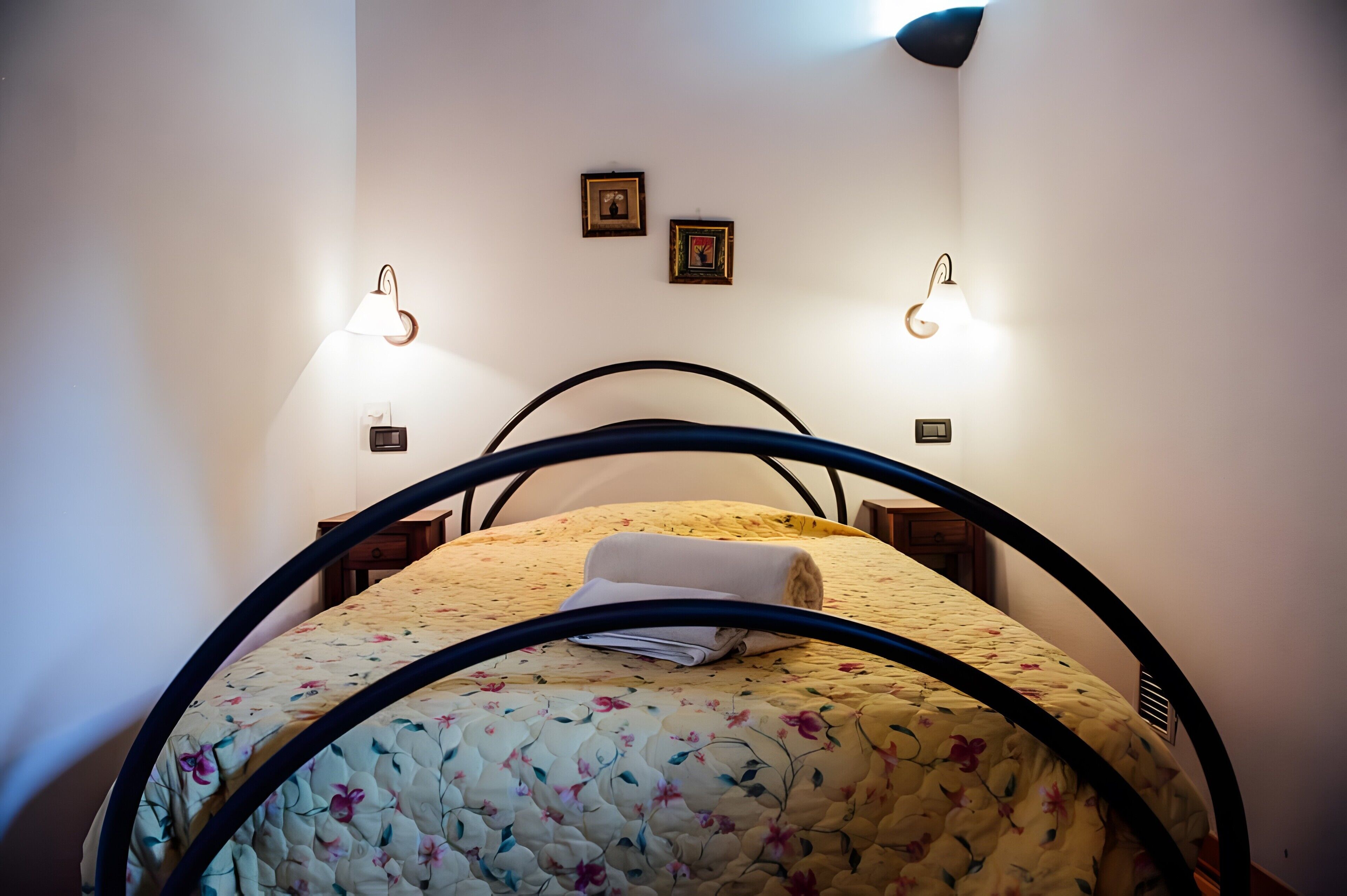 Un lit recouvert d’une courtepointe à motifs floraux, deux lampes fixées au mur et deux tableaux encadrés accrochés au mur.