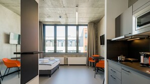 A modern hotel room with a bed, a desk, a chair, a kitchenette, and a large window offering a city view. - Nena Apartments Moritzplatz by LESA (Berlin)