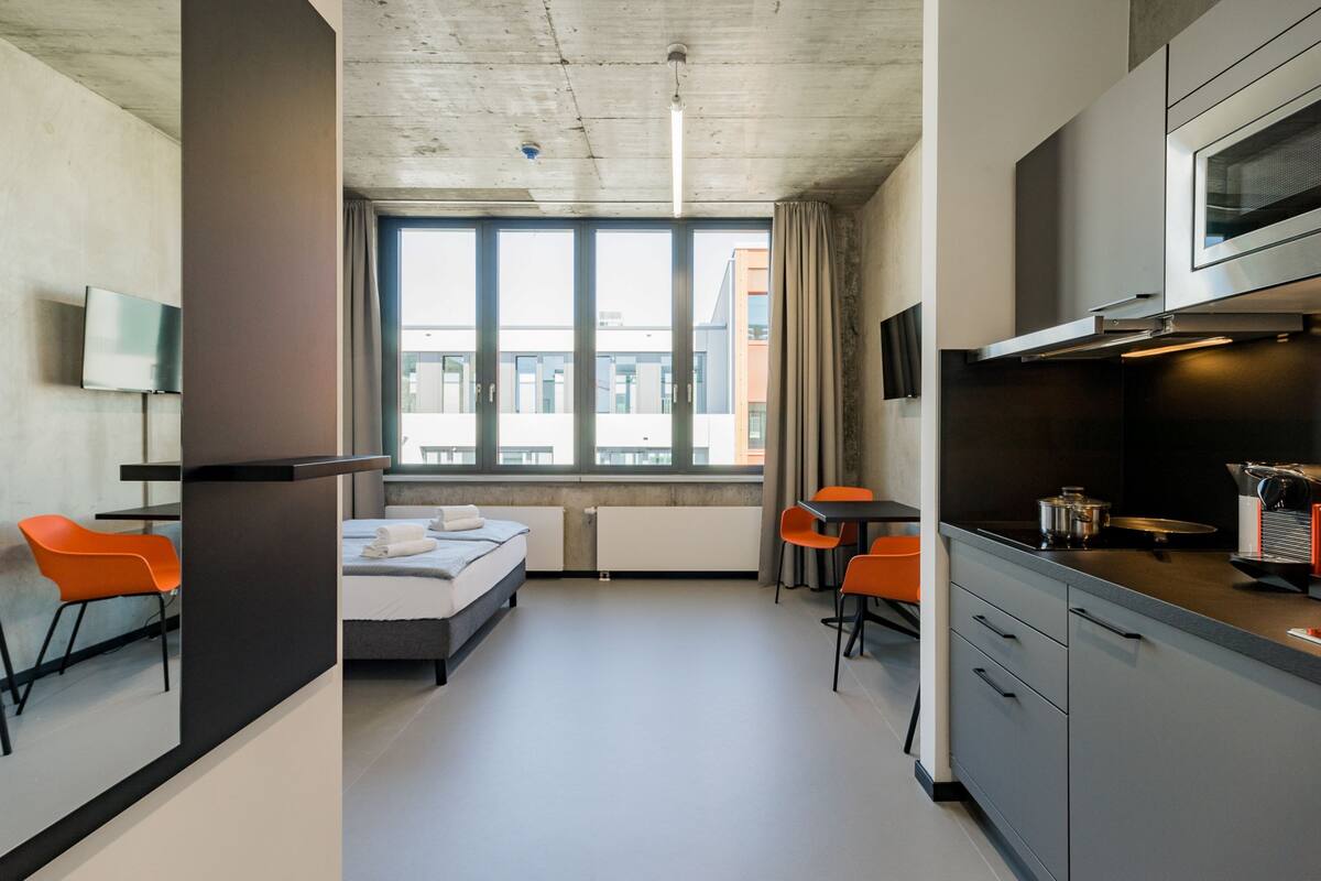 Ein modernes Hotelzimmer mit Bett, Schreibtisch, Stuhl, einer Küchenzeile und einem großen Fenster mit Blick auf die Stadt.