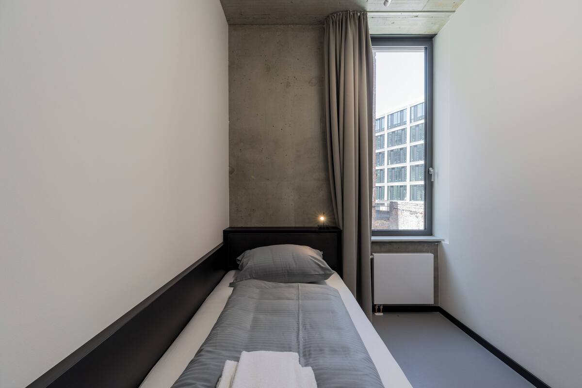 Ein kleines, modernes Hotelzimmer mit einem Einzelbett, einem Fenster mit Vorhängen, einer Betonwand und Blick auf ein Gebäude.