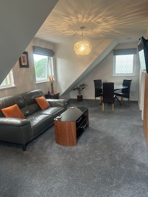 A modern loft-style room with a sloped ceiling, a leather sofa, a wooden coffee table, a dining table with chairs, and a potted plant.