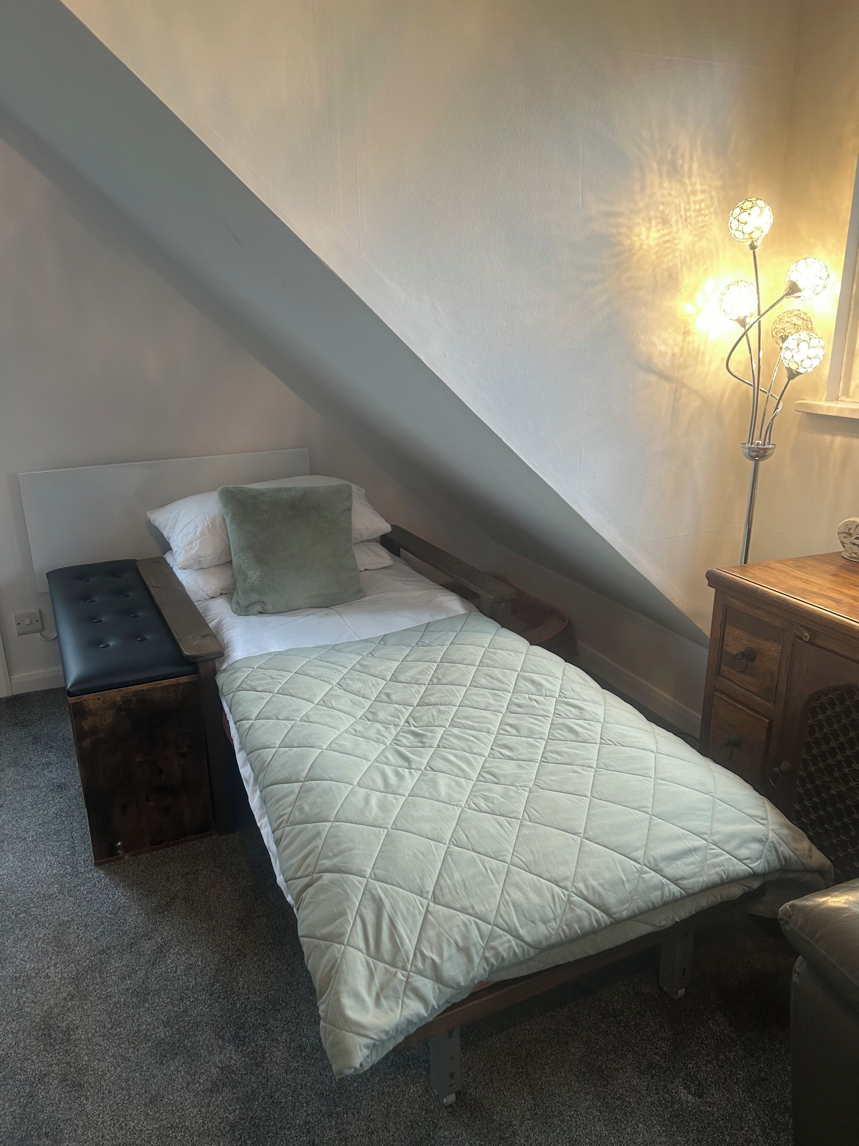 A small, neatly made bed with a quilted cover, a bedside bench, a nightstand, and a floor lamp with three bulbs.