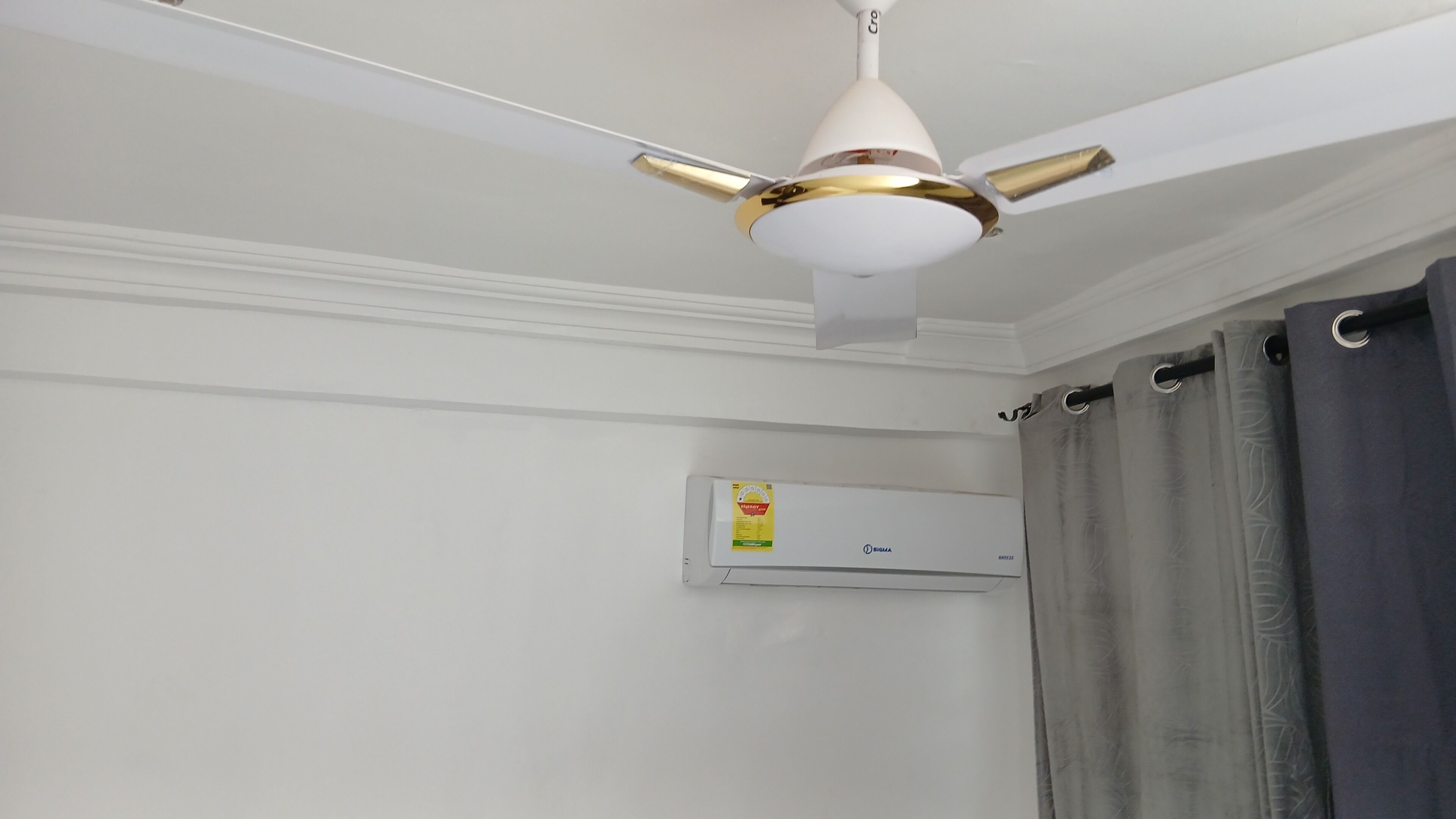 Ein Deckenventilator mit weiß-goldenem Design, eine Klimaanlage an der Wand und ein grauer Vorhang.