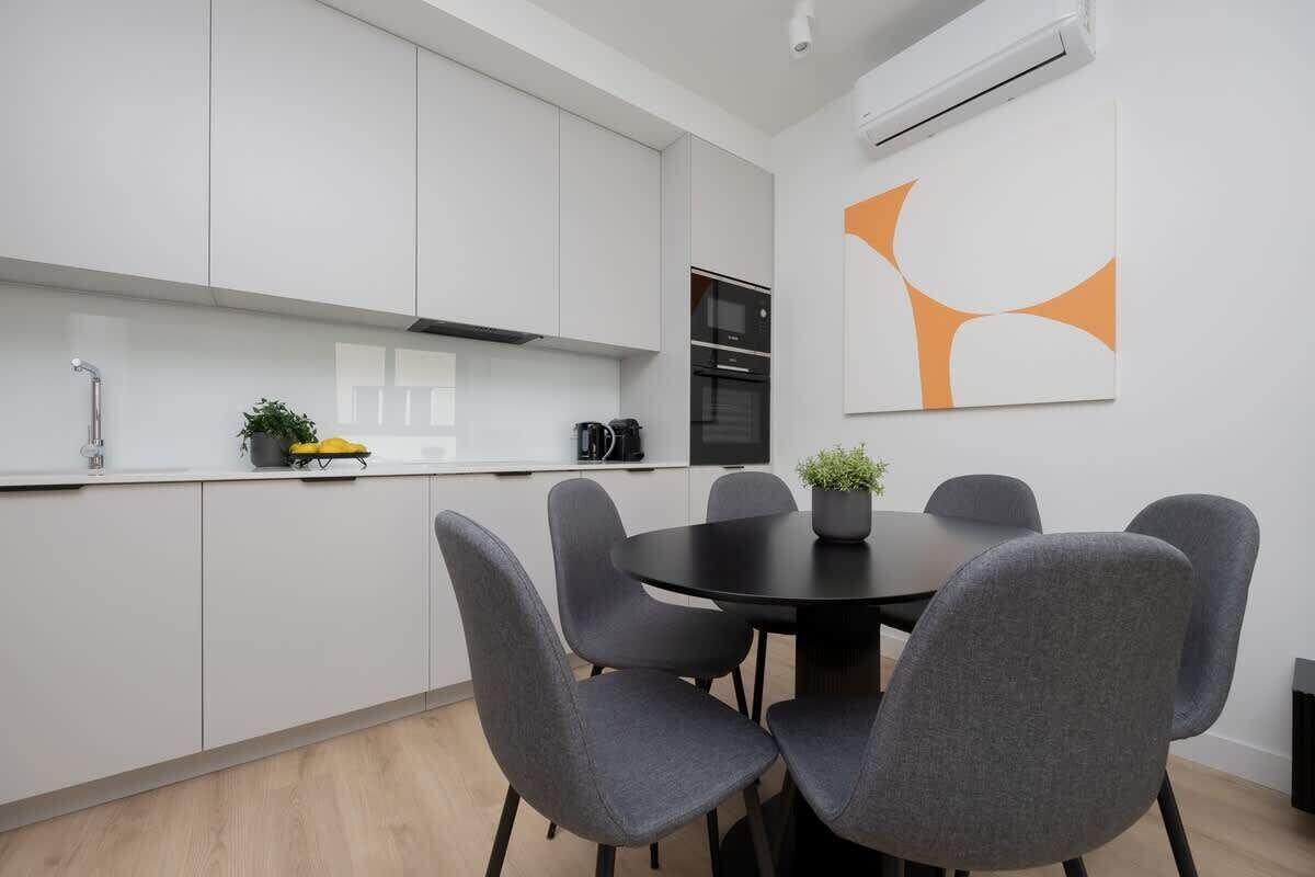A modern kitchen with white cabinets, a black stove, and a dining area with grey chairs.