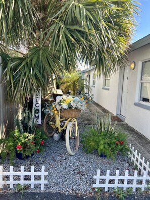 A bicycle with a basket of flowers and fruits parked under a tree, a white picket fence, and a 'WELCOME' sign. - Steps to Downtown, Dinner & Live Music (Palm Harbor)