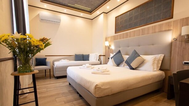 A hotel room with a large bed, a smaller bed, a desk, a chair, a vase with flowers, and a wall-mounted air conditioning unit. - Antica Piacenza (Piacenza)