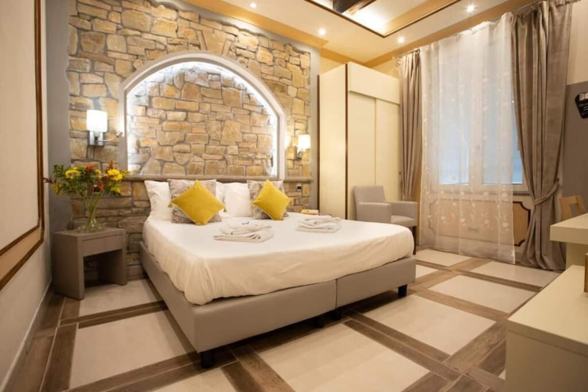 A bedroom with a stone wall, a bed with white bedding and yellow pillows, a bedside table, a chair, and a window with curtains.