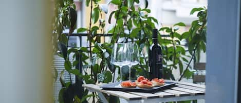 A table with a bottle, glasses, and food on a balcony with plants.