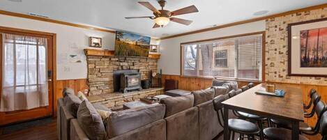 A cozy living room with a stone fireplace, a large sectional sofa, a dining table with bar stools, and a ceiling fan.