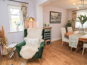 A room with a green armchair, a wooden dining table, a lamp, a framed picture on the wall, and a window with curtains. - The Coach House (Dorchester)