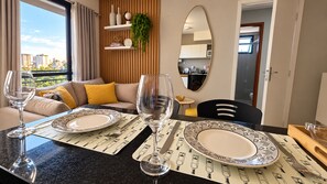 A dining area with a table set for two, a mirror, and a view of the cityscape through a window.