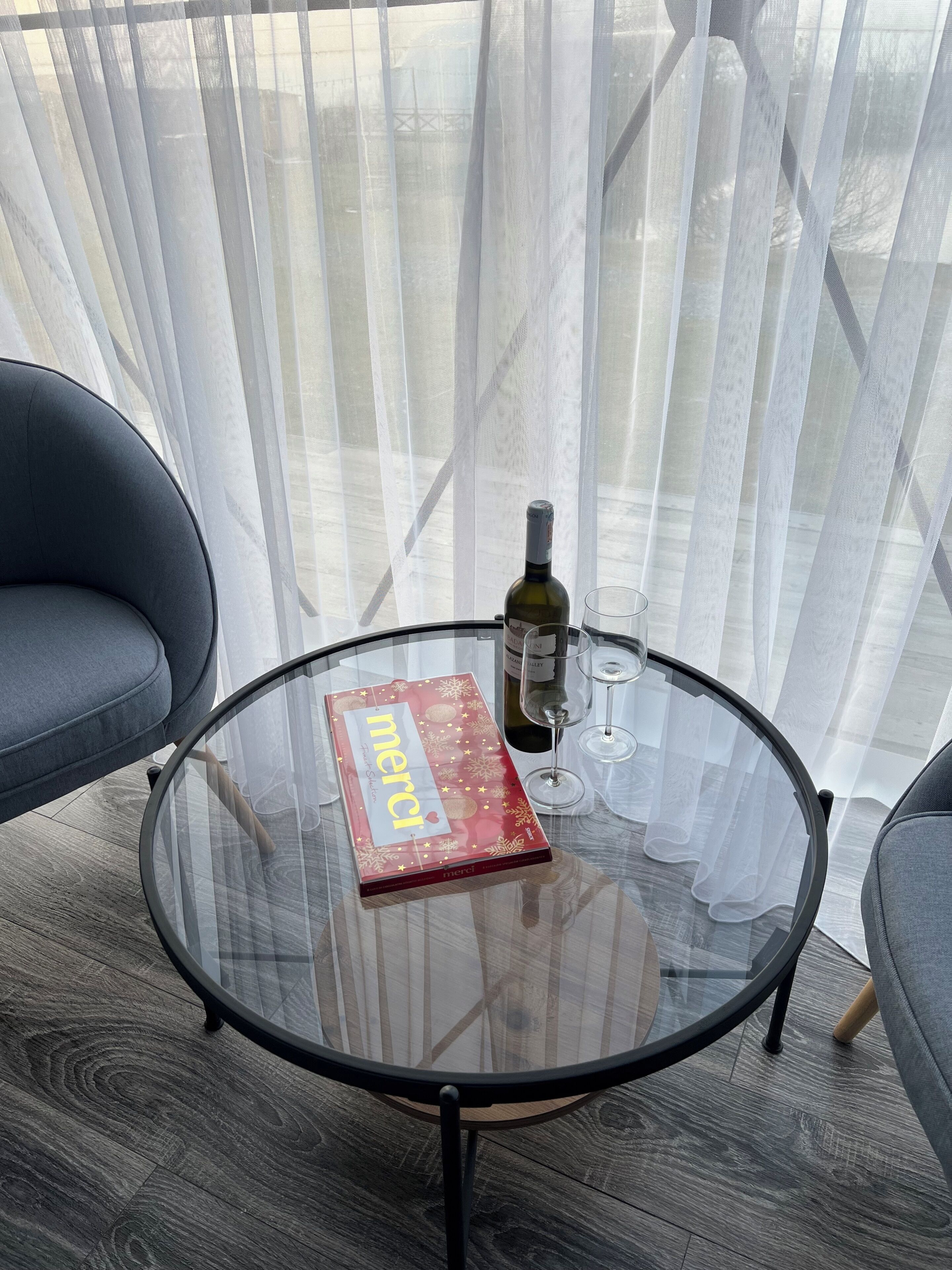 A round glass table with a book and a bottle, two grey chairs, and a sheer curtain backdrop.
