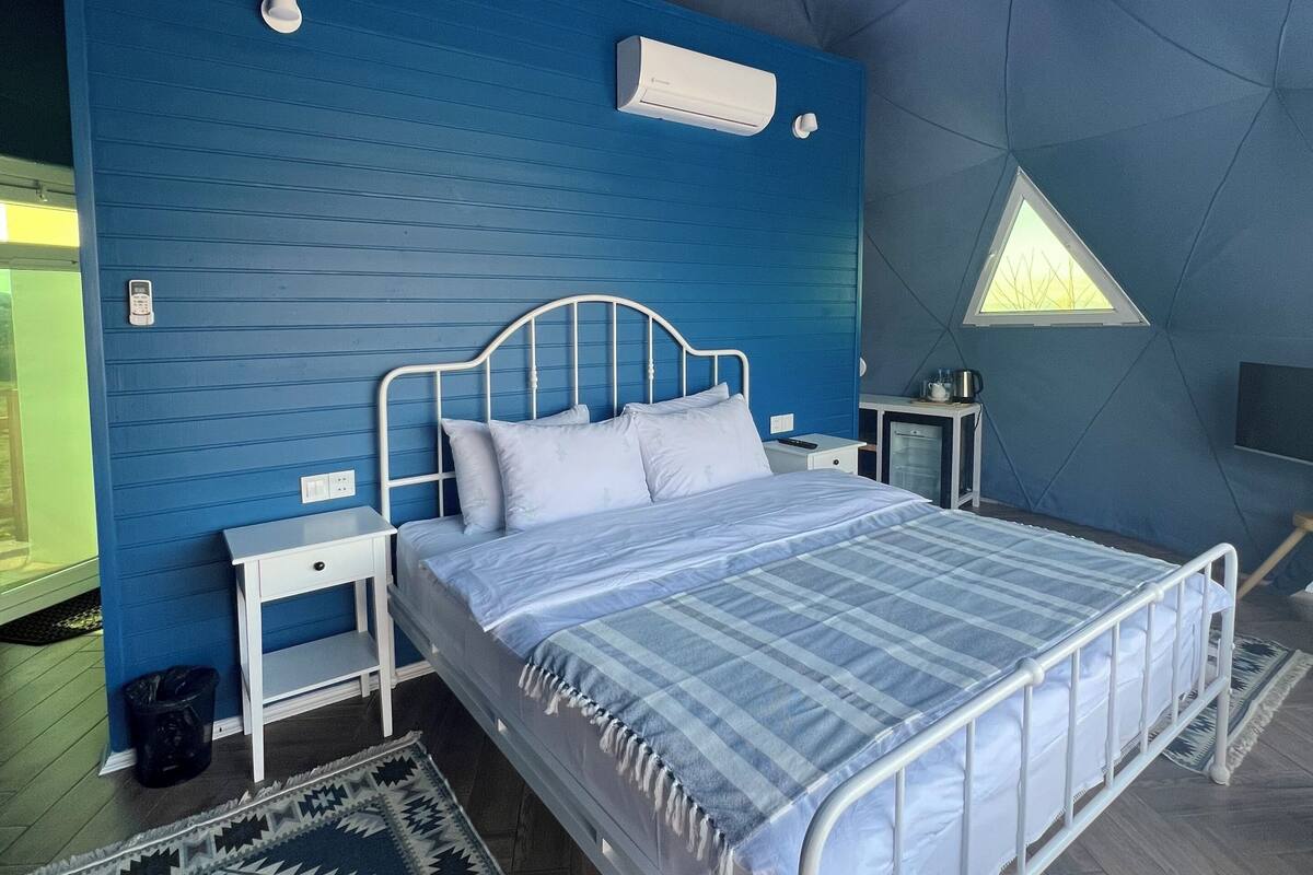 A bedroom with a bed, a nightstand, a bedside table, a chandelier, an air conditioning unit, a small window, and a patterned rug.