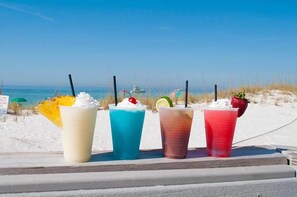 Vier farbenfrohe Getränke auf einem Tisch, im Hintergrund ein Strand und ein Ozean.