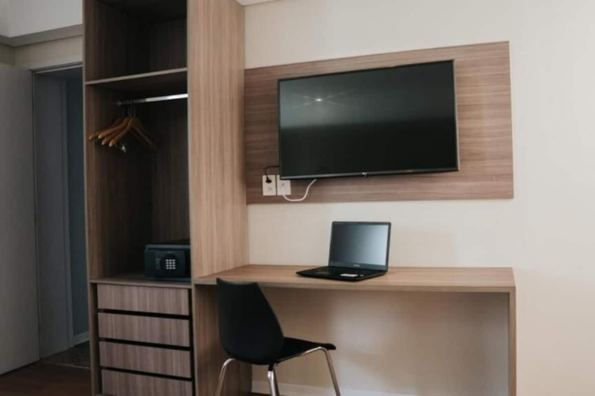 A modern room with a wooden desk, a black chair, a television mounted on the wall, and a wardrobe with hanging clothes.