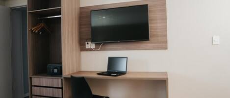 Una habitación moderna con un escritorio de madera, una silla negra, un televisor montado en la pared y un armario con ropa colgada.