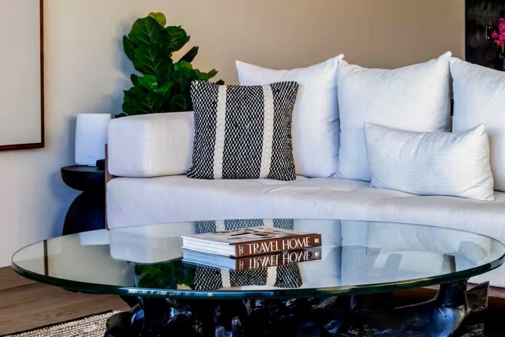 Uma sala de estar moderna com uma mesa de centro de vidro, um sofá branco e uma almofada decorativa estampada.