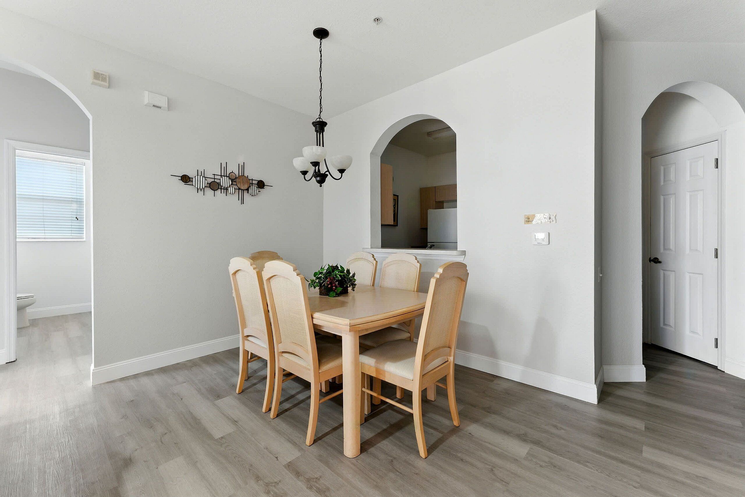 Un espace repas avec une table et des chaises en bois, un lustre et une cuisine visible à travers une arche.